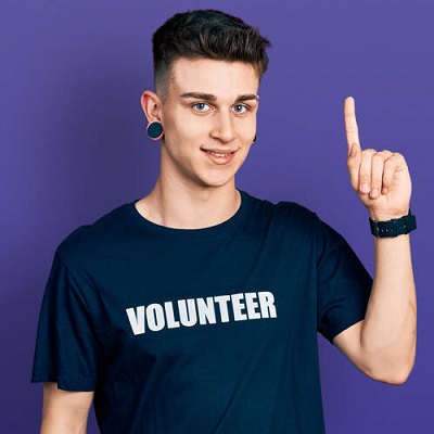 Young caucasian boy with ears dilation wearing volunteer t shirt with a big smile on face, pointing with hand finger to the side looking at the camera.